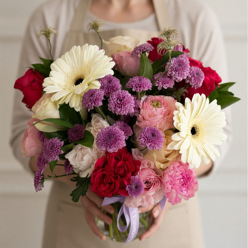 A person in a beige apron holds a large, vibrant flower bouquet featuring white Gerbera daisies, pink and red roses, purple chrysanthemums, and green foliage.