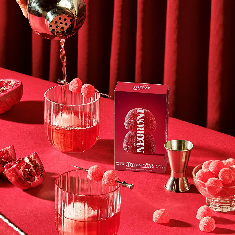 Negroni gummies being poured into glasses on a red table with Negroni Gummies packaging.