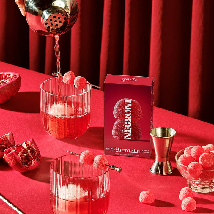 Negroni gummies being poured into glasses on a red table with Negroni Gummies packaging.
