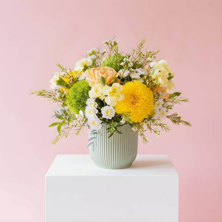 A floral arrangement in a ribbed pale green vase, placed on a white pedestal against a soft pink background. The bouquet includes yellow, white, peach, and green flowers with mixed green foliage.