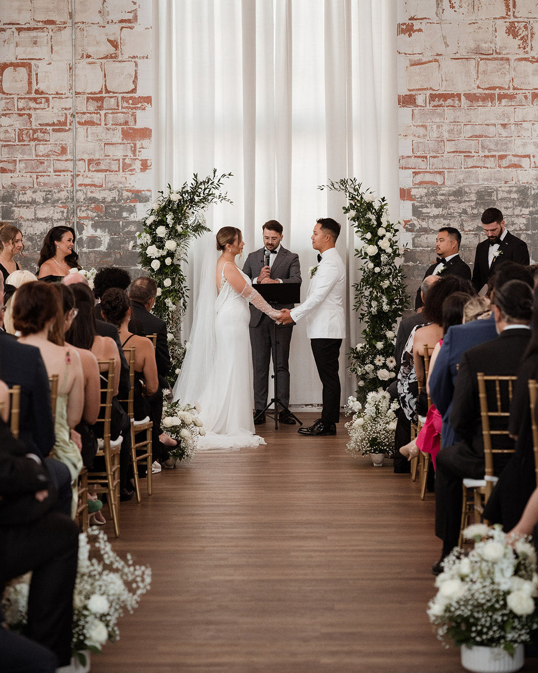 Wedding ceremony taking place in a room with brick walls and white curtains.