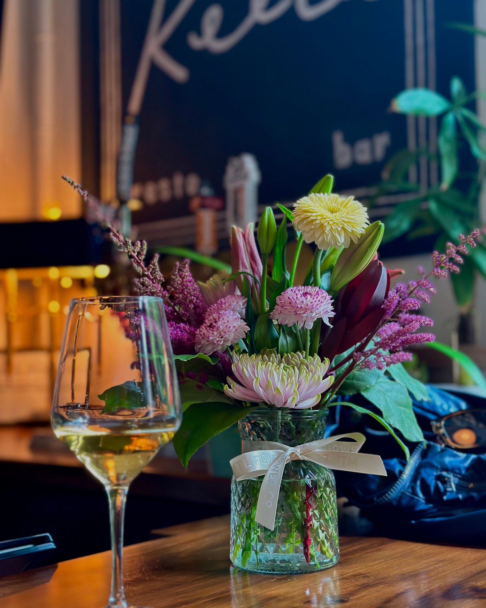 Floral arrangement in a vase with a wine glass on a table, blurred background with 'Keego' bar sign.