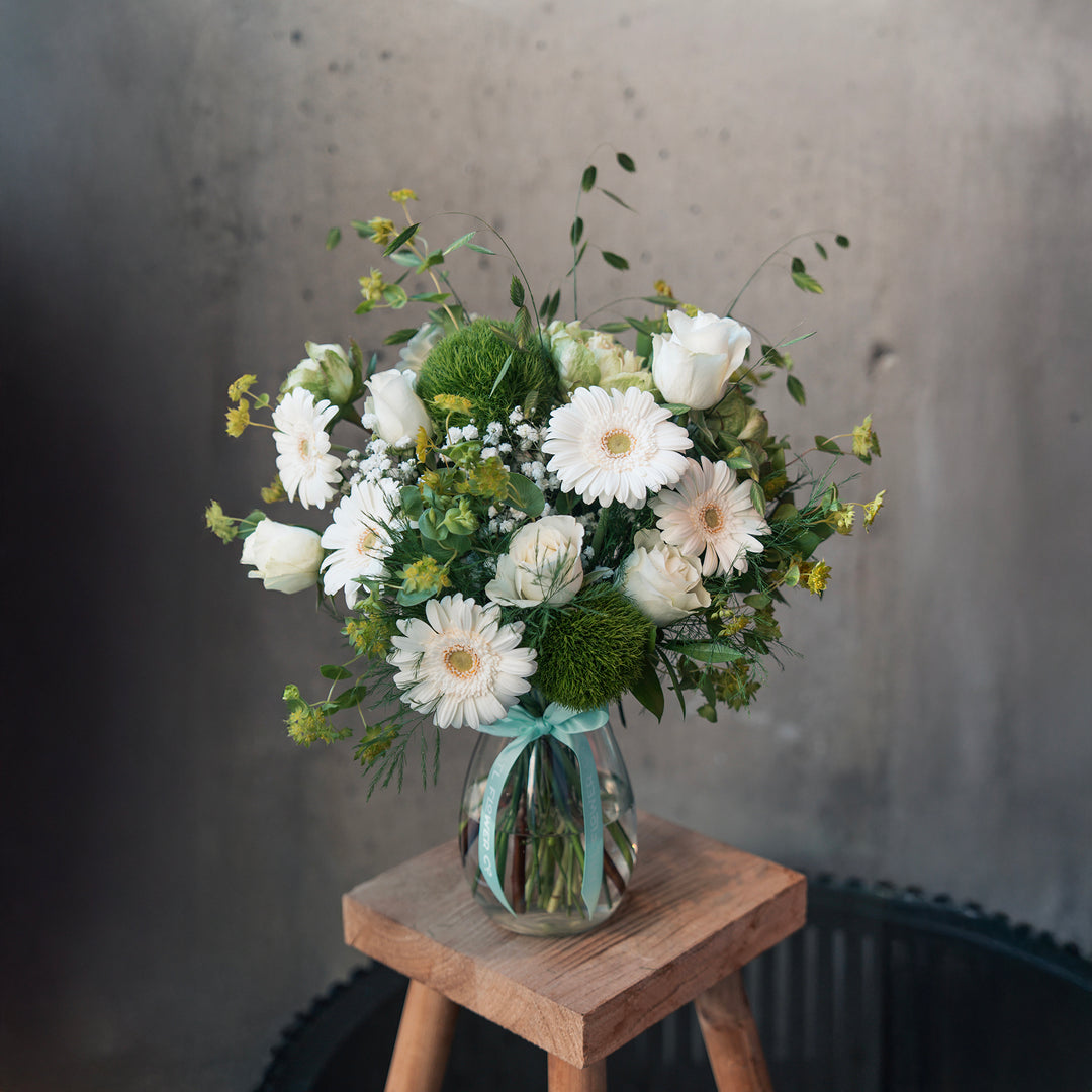 Bouquet of white and green flowers in a clear vase on a wooden stool against a gray background