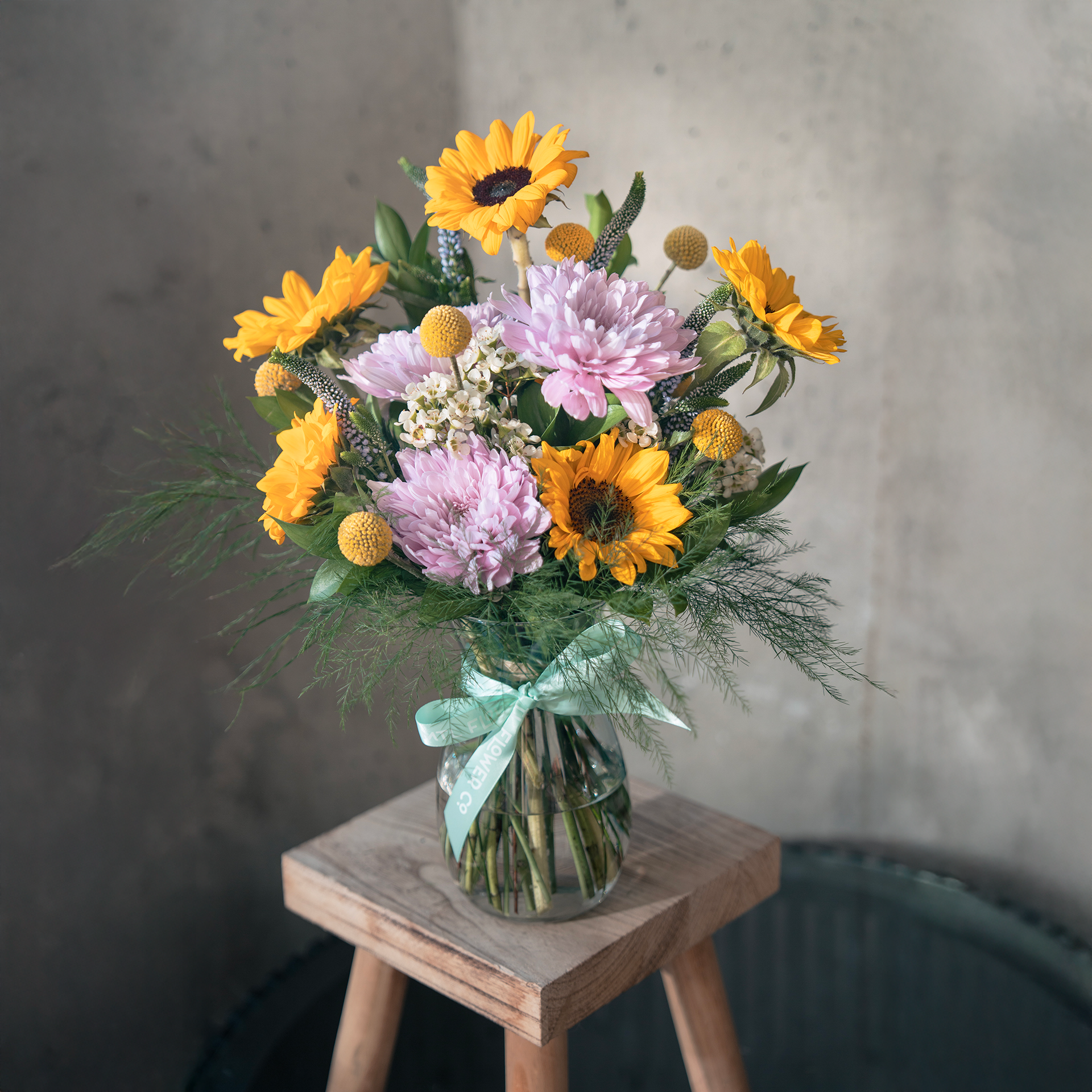 Bouquet of sunflowers and pink flowers in a clear vase on a wooden stool with a neutral background
