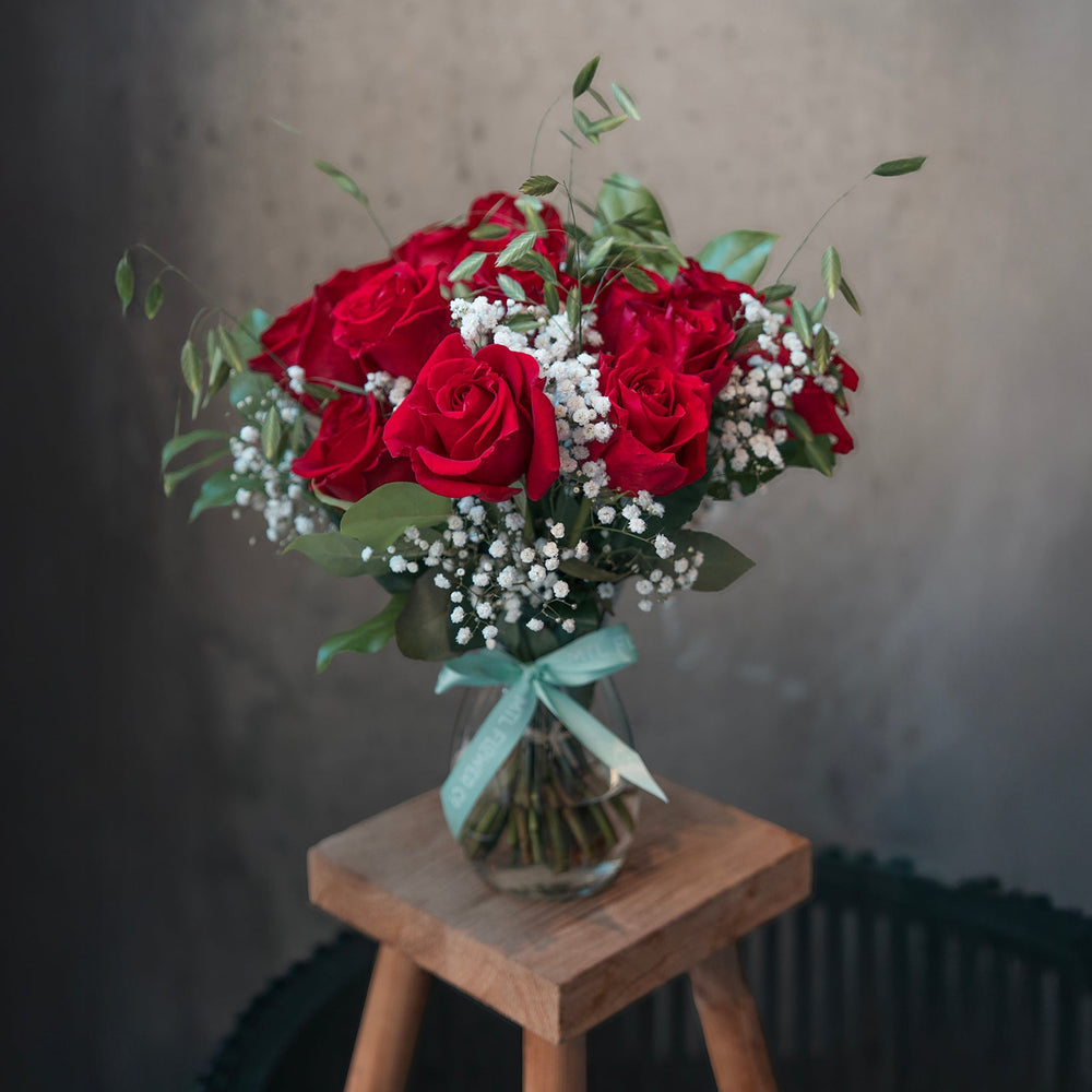 Bouquet of red roses with greenery and white flowers in a clear vase on a wooden stool against a neutral background.