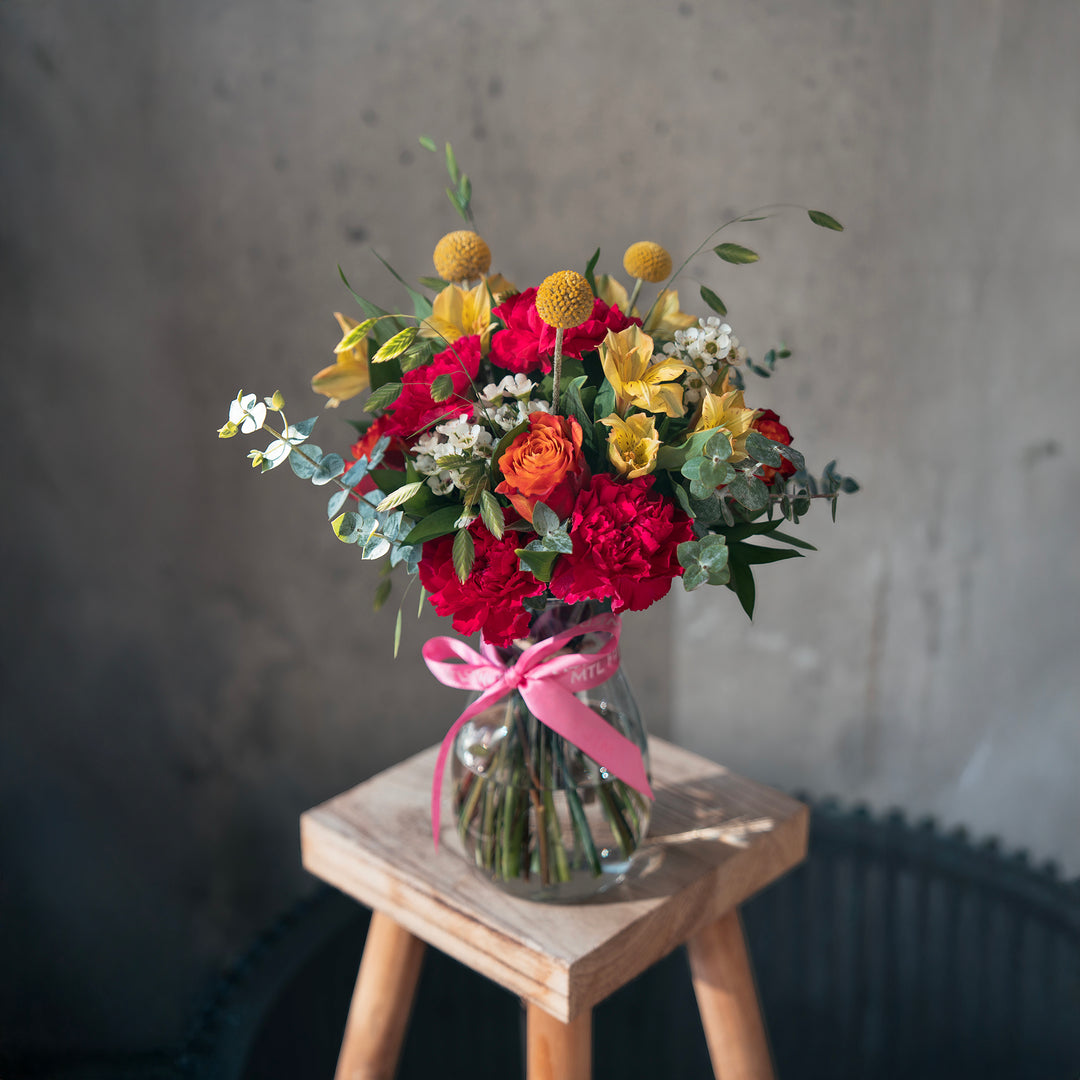 Bouquet of flowers in a glass vase with a pink ribbon on a wooden stool against a gray wall.