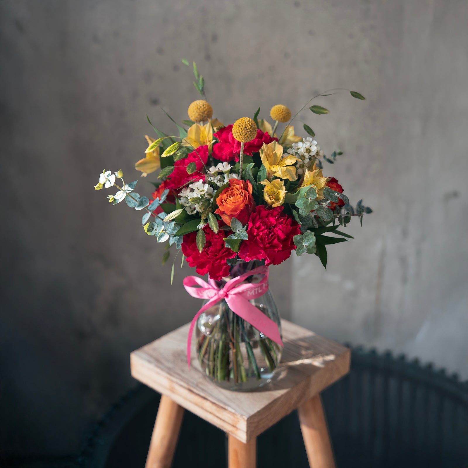 Bouquet of flowers in a glass vase with a pink ribbon on a wooden stool against a gray wall.