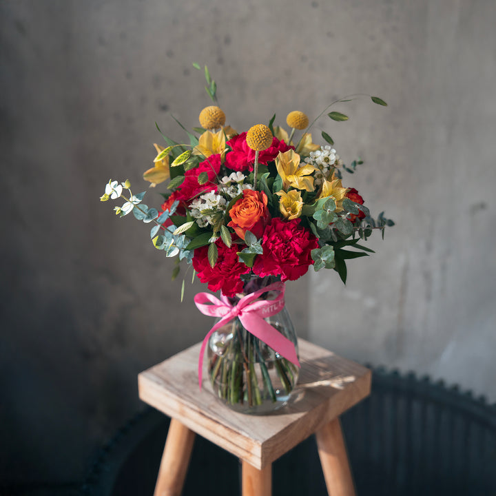Bouquet of flowers in a glass vase with a pink ribbon on a wooden stool against a gray wall.
