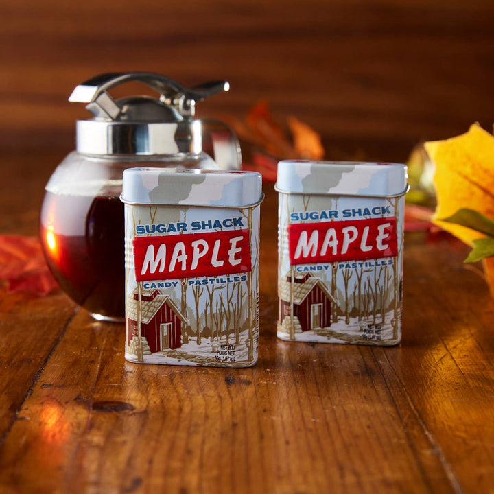 Two containers of Sugar Shack Maple syrup on a wooden surface with autumn leaves.