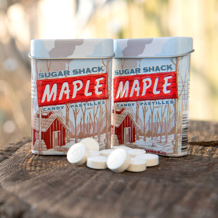 Two tins labeled 'Sugar Shack Maple' on a wooden log with a blurred natural background.